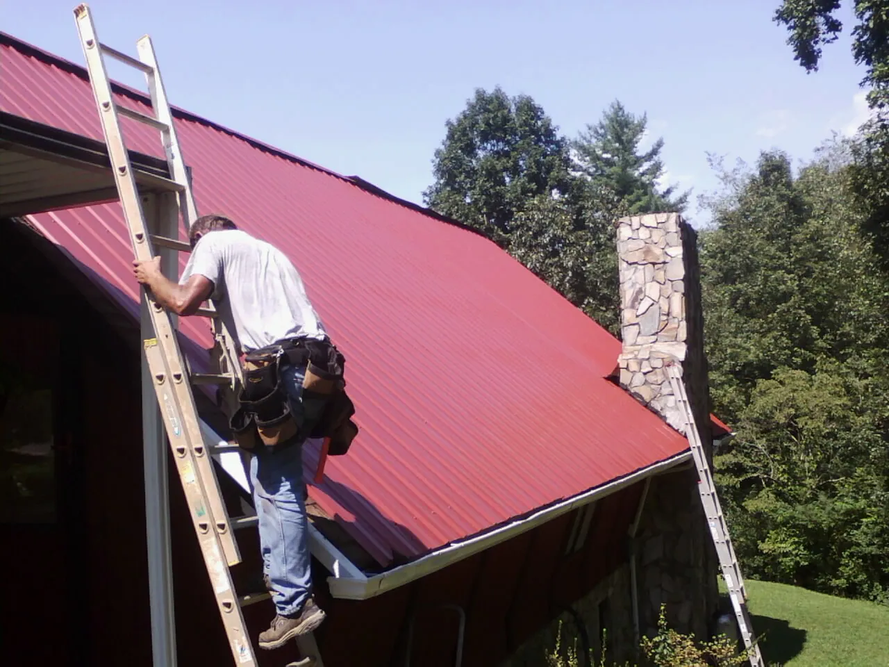 Roofer on a ladder beside a red metal roof and stone chimney — Soffit & Fascia Repair by Roofing Brothers in Waterford Works